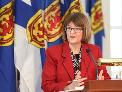 Diana Whalen at a press conference—she is standing at a podium, behind her are several Nova Scotia flags.