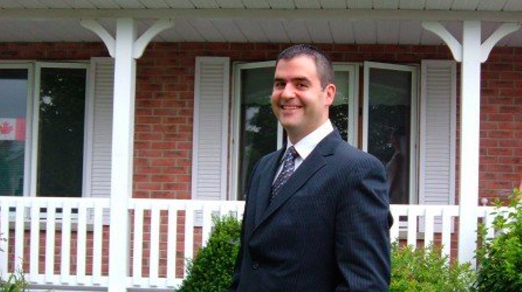 Aaron MacKenzie, a part-time teacher and full-time believer in trickle-up economics, stands in a suit in front of his house.